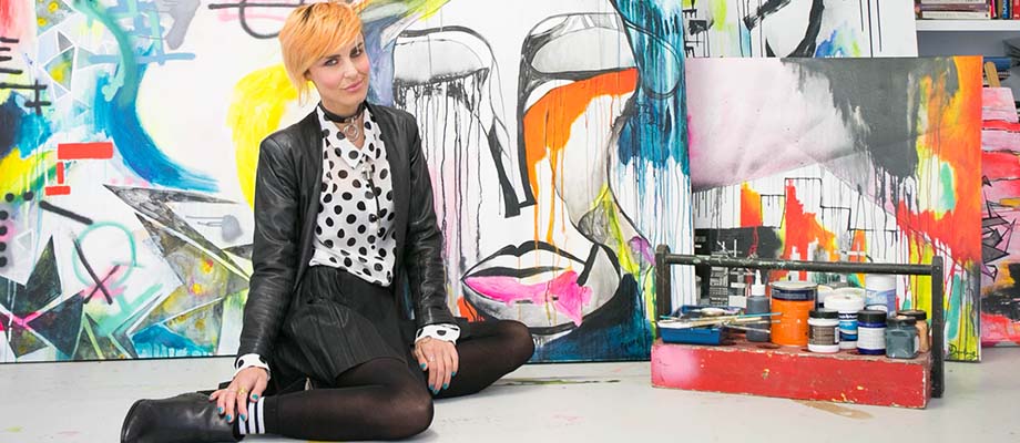 Woman sits on floor next to painting supplies, with artwork displayed behind and beside her