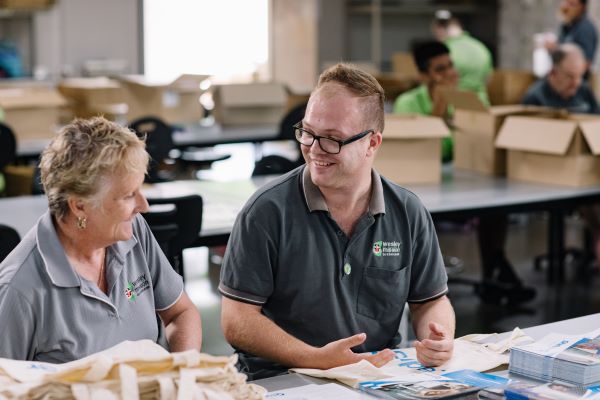 Two Wesley Industries workers sit side by side, laughing, with bags and pamphlets on table in front of them