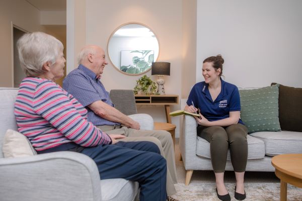 Two people sitting and talking to staff member with notebook in home environment