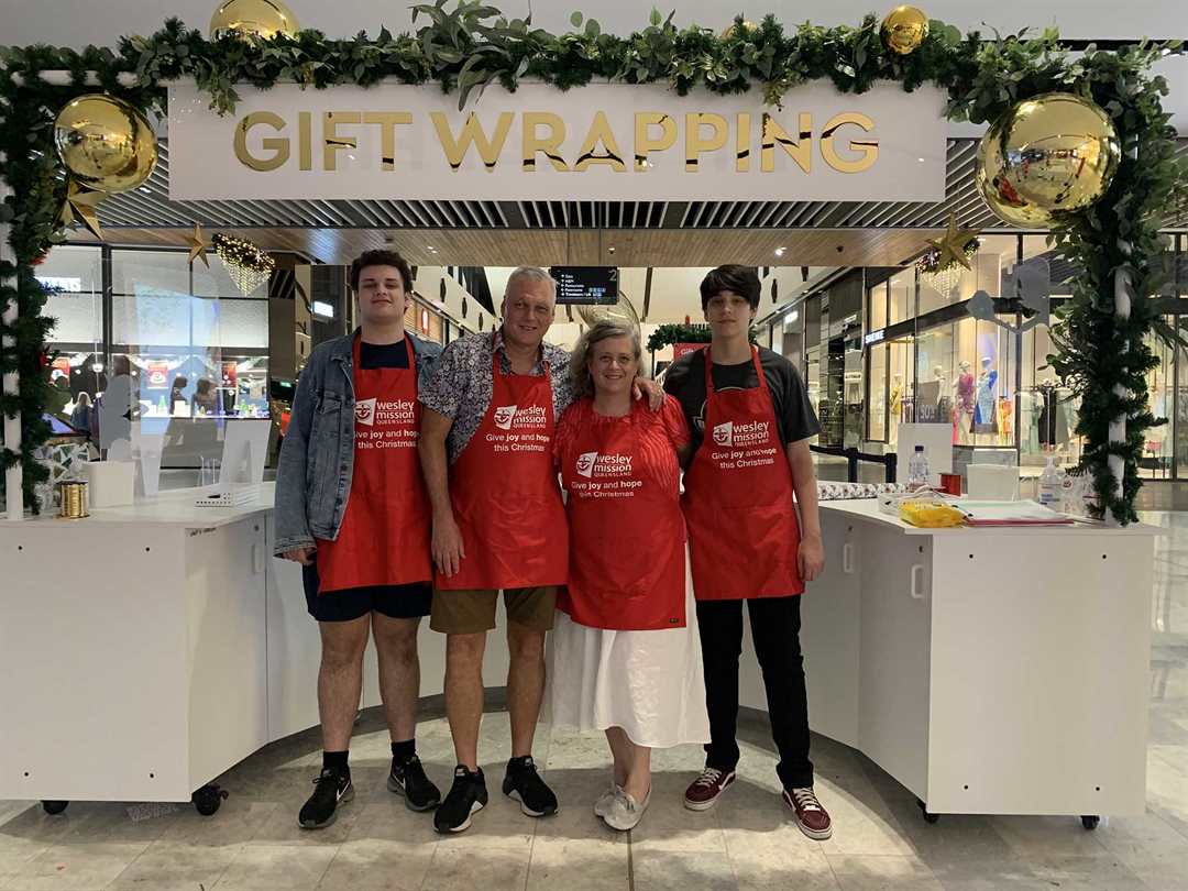 Four people wearing red aprons stand in front of stall with sign that says 'Gift Wrapping'