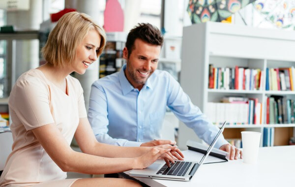A woman and man in professional clothing sit smiling and looking at laptop