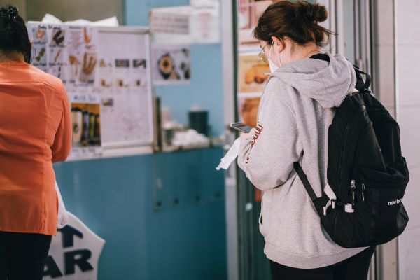 Person stands in line behind desk, wearing backpack and face mask and looking at phone