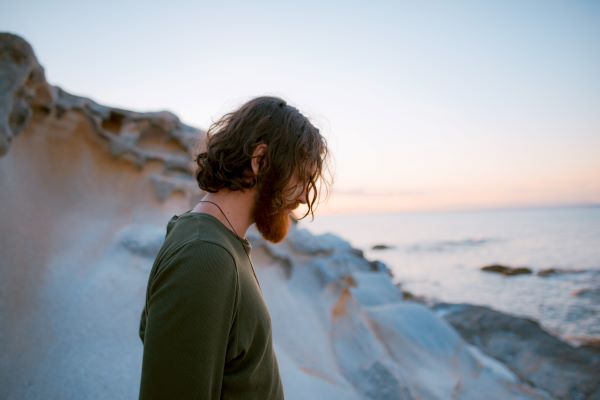 Man with long hair and beard stands on cliff and looks down at water