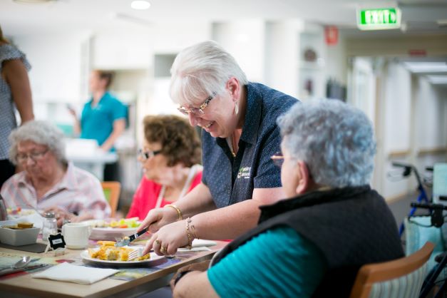 Staff member assisting a resident by cutting up their meal at Jacob's Court residential aged care home Social connections during mealtime at Jacob's Court residential aged care home