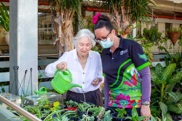 Older lady watering plants while masked staff member looks on