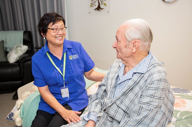 A care worker and elderly gentlemen sitting on bed smiling at each other