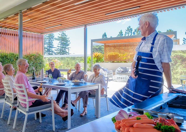 Man cooks on BBQ while talking to friends seated at table nearby