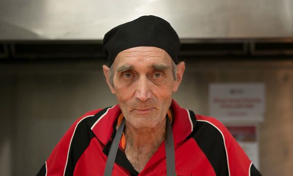 Man wearing apron and kitchen hat, gently smiling at camera.