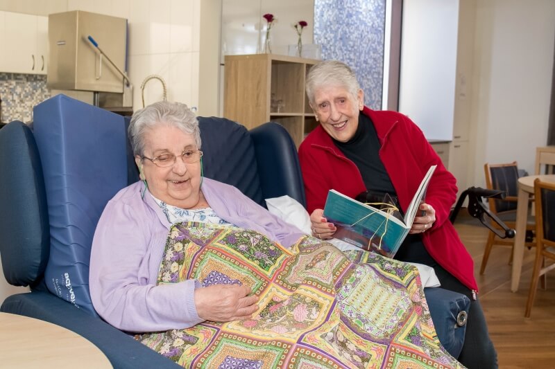 Two elderly ladies smiling. One is in a red jacket holding a book she is reading to the other in a purple cardigan with a crochet blanket over her
