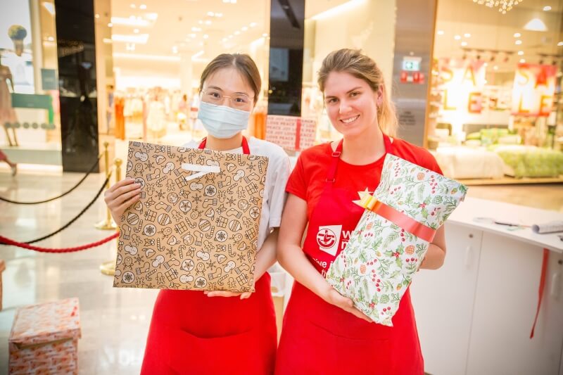 Two women wearing red aprons holding presents wrapped in Christmas paper and smiling