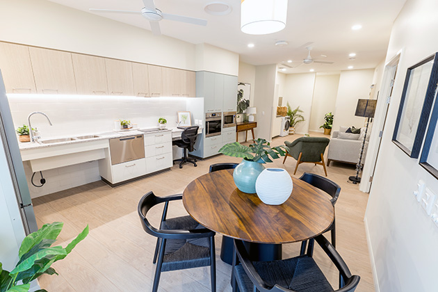 A round dining table with a vase of flowers on it is pictured in the kitchen/dining area of a supported disability accommodation home with the living roomw and two couches in the background