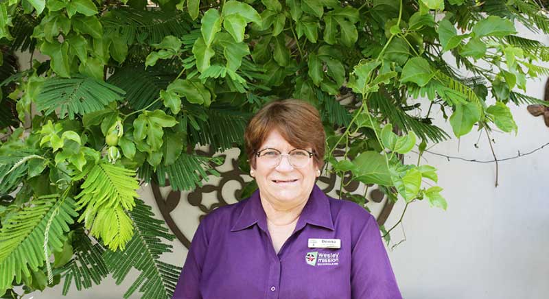 Lady in Wesley Mission Queensland uniform smiles at camera with plants behind