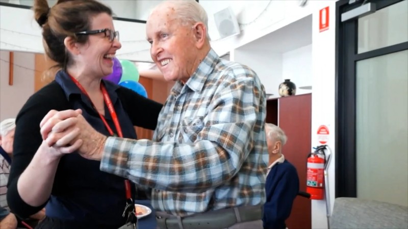 Woman in a black jumper wearing glasses laughing as she holds an older man in a check shirt's hand and dances with him