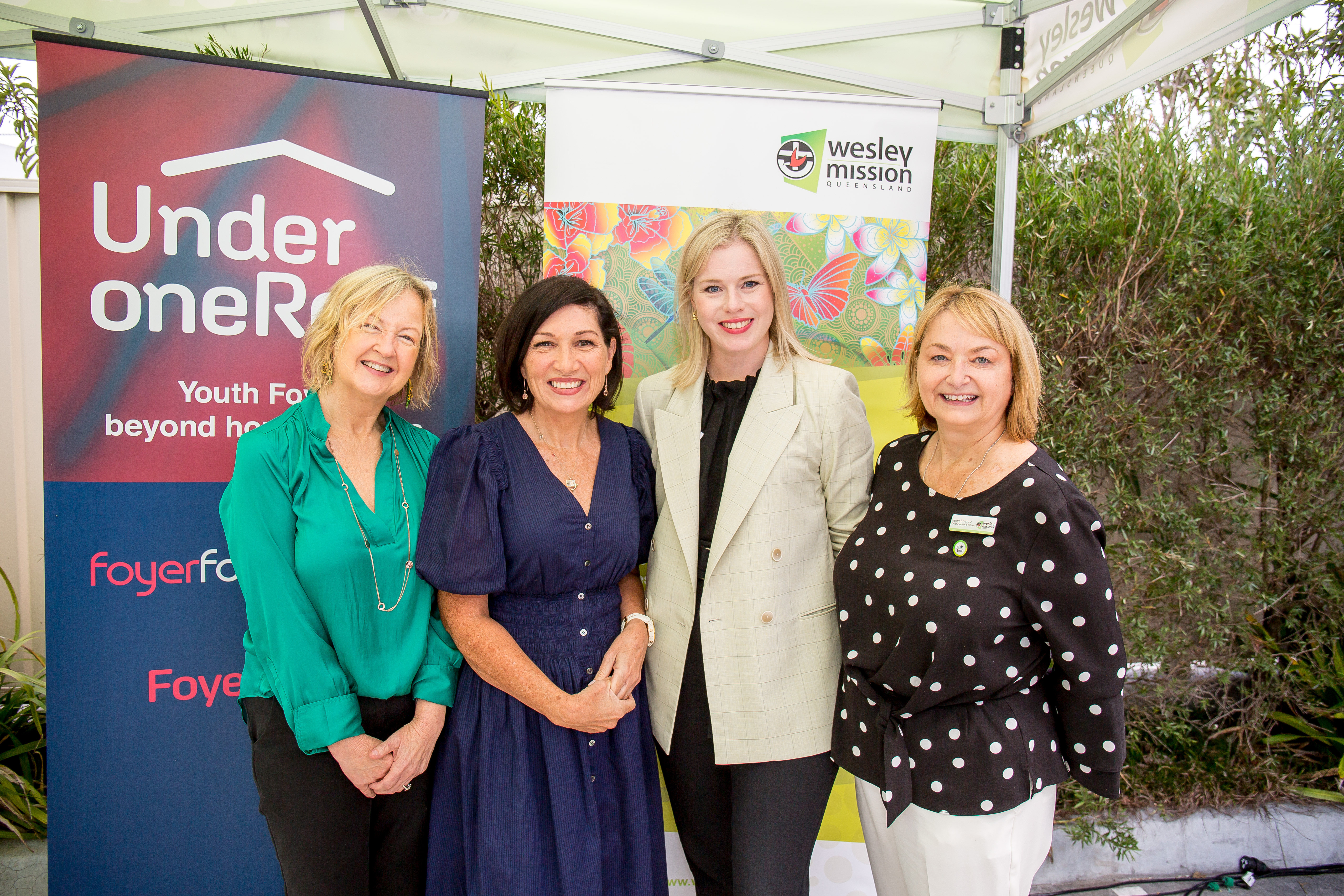 Four middle aged women standing in front of two pull banners outside with green shrubs in the background