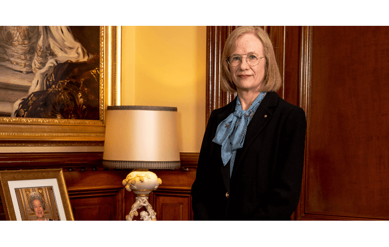 Queensland Governor Dr Jeanette Young standing dressed in a suit jacket in front of a photo of Queen Elizabeth II