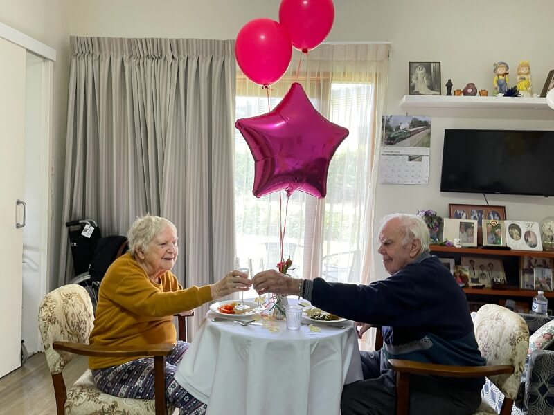 Elderly man and woman sitting at a table with a white table cloth and balloons in the background clinking champagne glasses