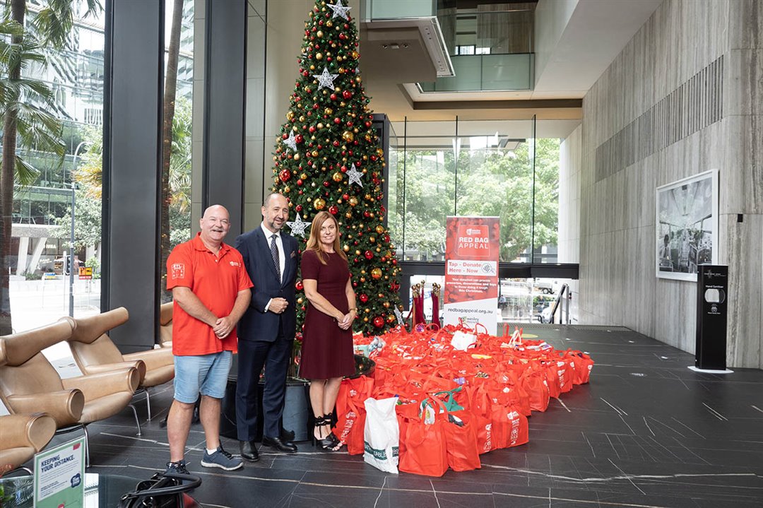 Three people stand beside Christmas tree and many red grocery bags