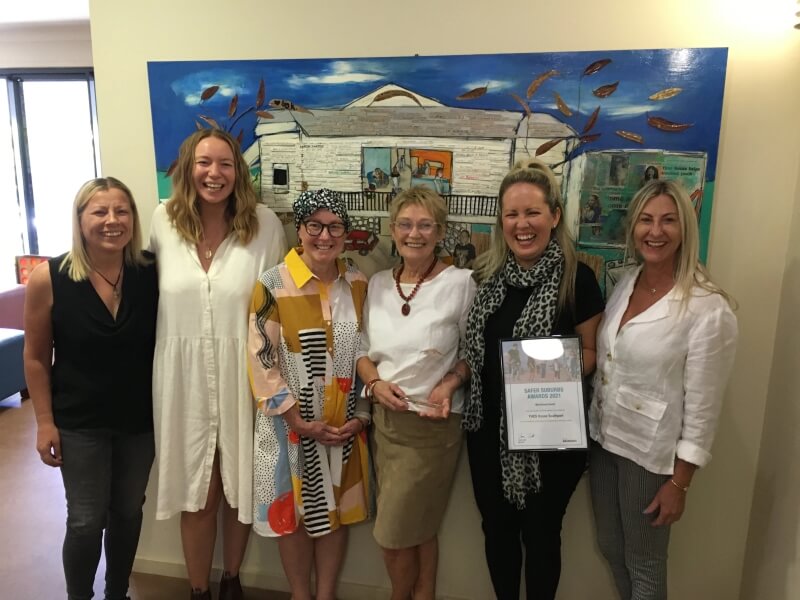 Group of six women standing in front of a painting holding an award and looking happy