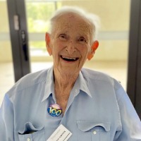 Elderly man wearing a blue collared shirt and a badge saying 100 smiling